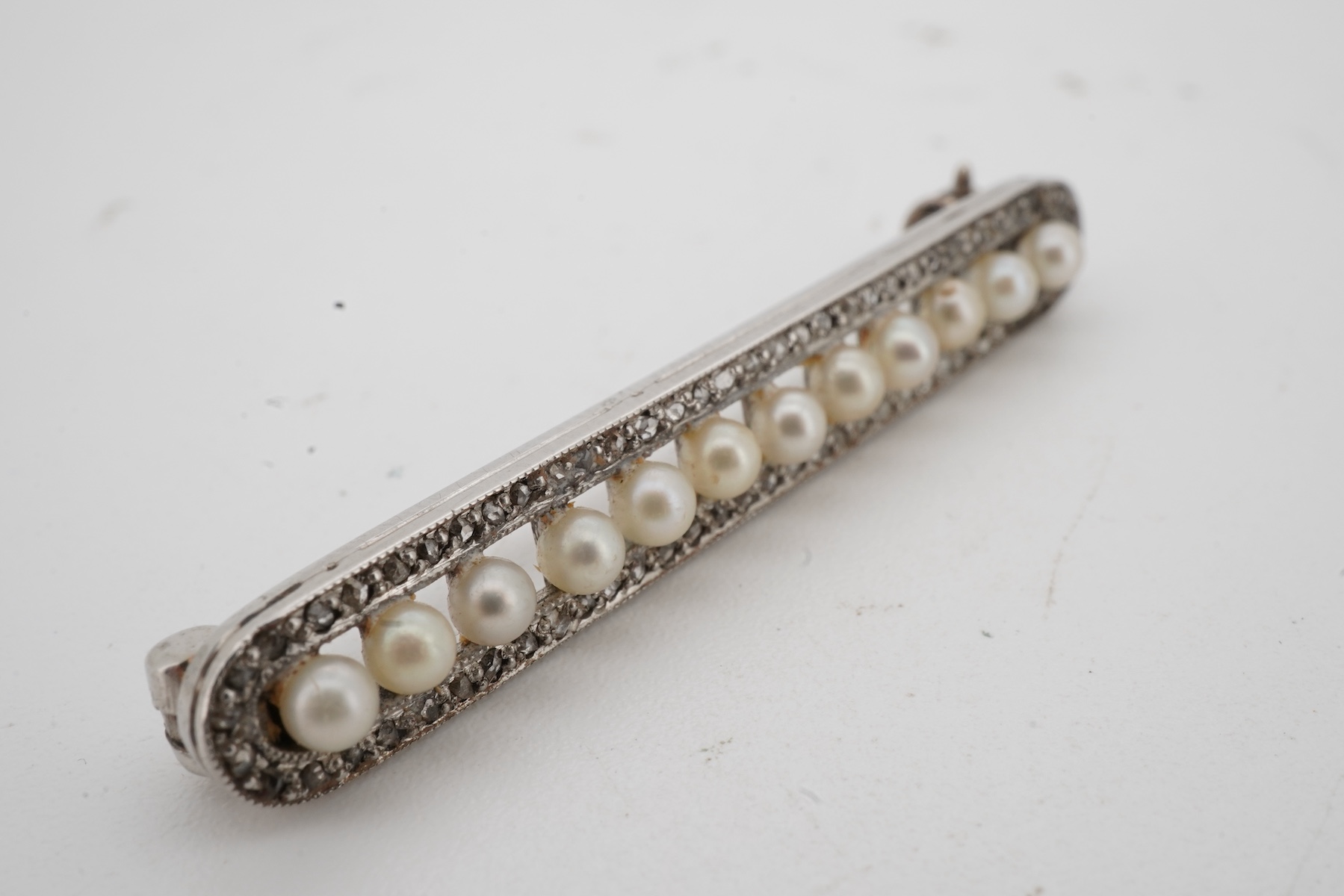 A Belle Epoque pearl and diamond brooch, early 20th century, of bar design, composed of a line of seed pearls within an oval border of rose-cut diamonds, mounted in platinum, length 3.9cm, French sponsor's mark, partial